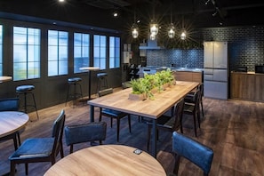 A modern interior with a long wooden table, bar stools, and a kitchen area with a refrigerator. - ALPHABED INN Otaru Ekimae (Otaru-shi)