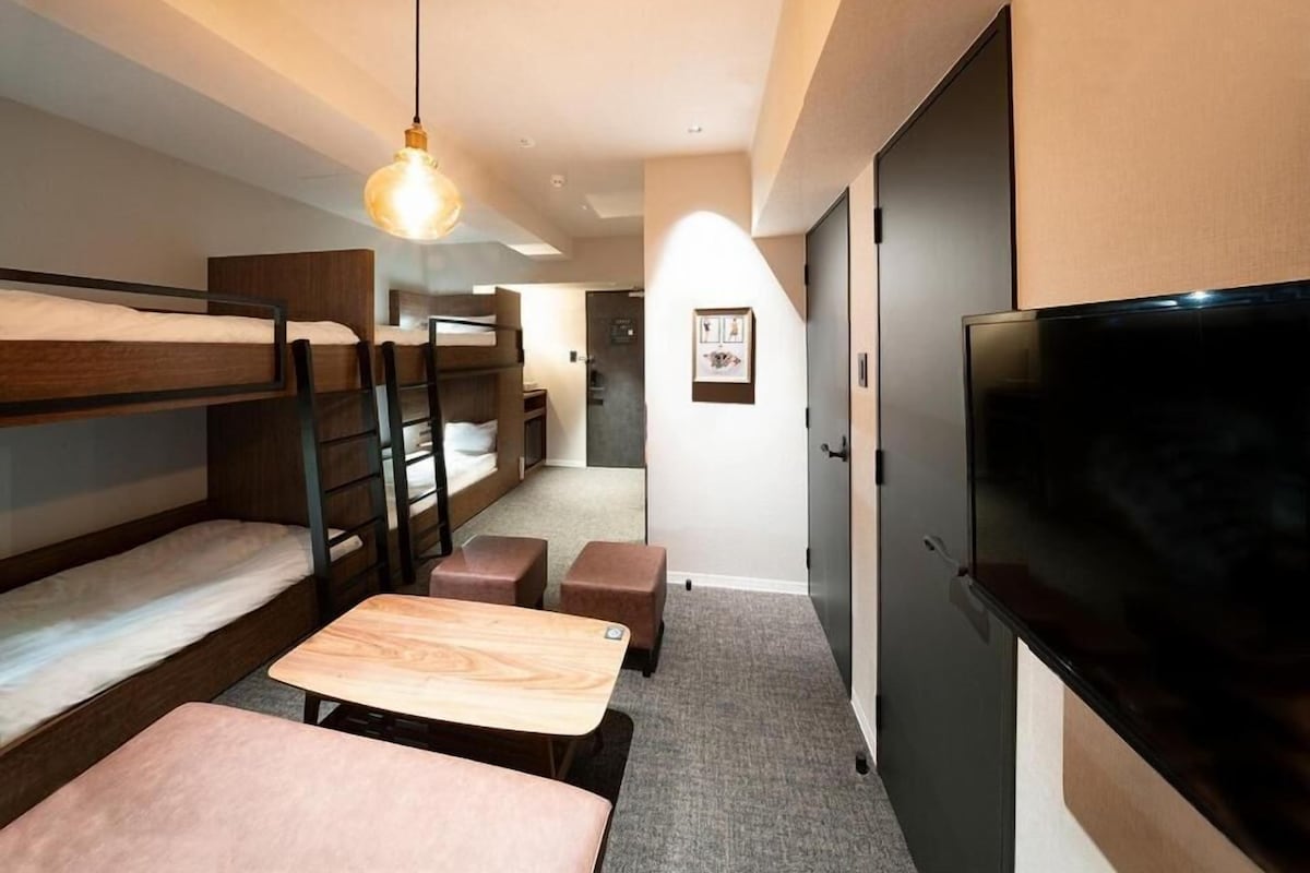 A compact hotel room with bunk beds, a flat-screen TV, a wooden table, and a hanging light fixture.