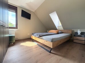 Ein Schlafzimmer mit schrägem Dach, einem Bett mit Holzrahmen, einem Nachttisch und einem Fenster.