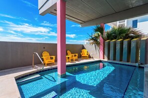 Une piscine à l’eau d’un bleu limpide, entourée d’une terrasse en béton et agrémentée d’une colonne rose. On y trouve des chaises jaunes et une petite table près de la piscine.