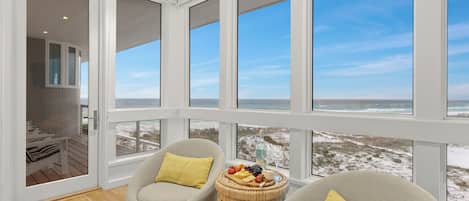 Una habitación con ventanas amplias que dan a una playa, amoblada con dos sillas y una mesita con aperitivos.