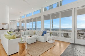 Un salon spacieux avec un canapé blanc, une table basse et une vue sur l’océan grâce à de grandes fenêtres.