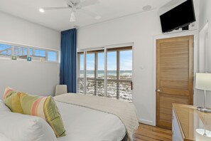 A bedroom with a bed, a television, a window with a view of the beach, and a door with a wooden panel. - Gulf-Front Retreat • Pool + Golf Cart • Near Gulf Place | Matarangi (Santa Rosa Beach)