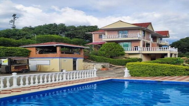 Une piscine bordée d’une balustrade blanche, un bâtiment à deux étages doté de balcons, et un paysage verdoyant luxuriant en arrière-plan.