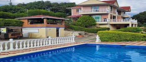 Una piscina con barandilla blanca, un edificio de dos pisos con balcones y un paisaje verde exuberante al fondo.