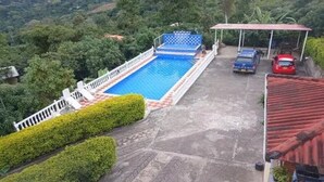 Une piscine entourée d’une clôture blanche et dotée d’un espace abrité, le tout bordé de verdure et d’une zone pavée où sont garées des voitures.