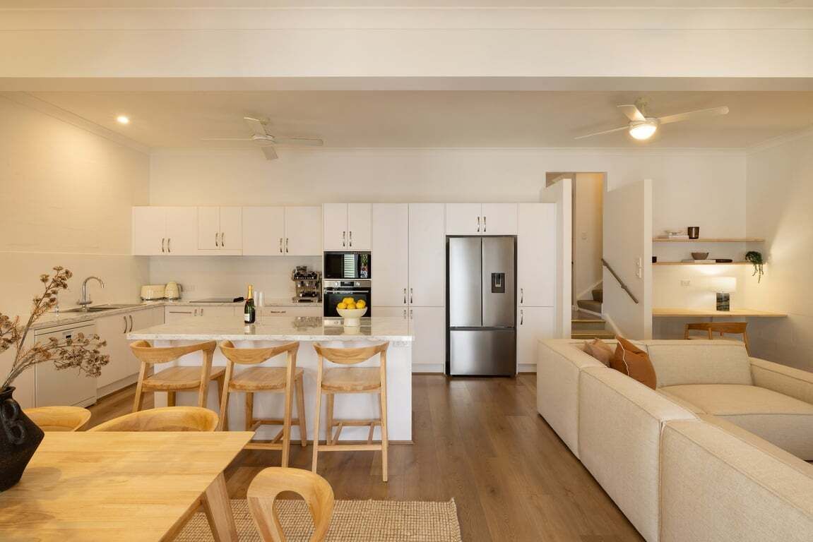 A modern kitchen with white cabinetry, a central island with bar stools, a stainless steel refrigerator, and a dining area with a wooden table and chairs.