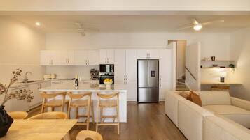 A modern kitchen with white cabinetry, a central island with bar stools, a stainless steel refrigerator, and a dining area with a wooden table and chairs.