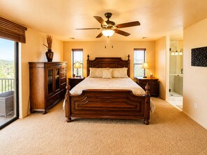 Un dormitorio con una cama grande, ventilador de techo, mesita de noche con lámpara, cómoda y vistas al exterior.