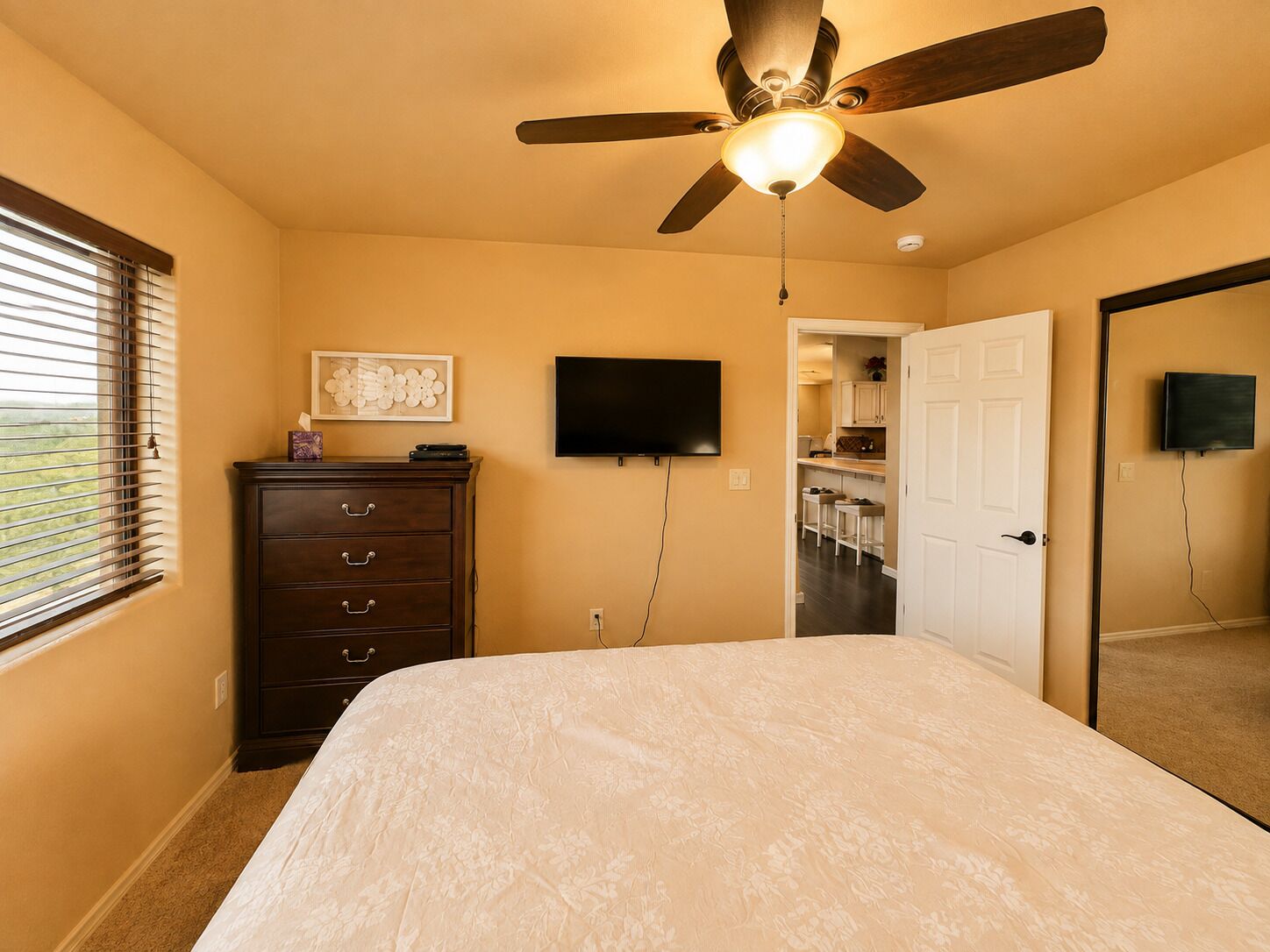 A bedroom with a bed, dresser, ceiling fan, TV, and a door leading to a kitchen.