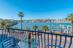 Un balcon avec une table, des chaises et une bouteille de vin, donnant sur une marina où sont amarrés des bateaux et sur une ville côtière.