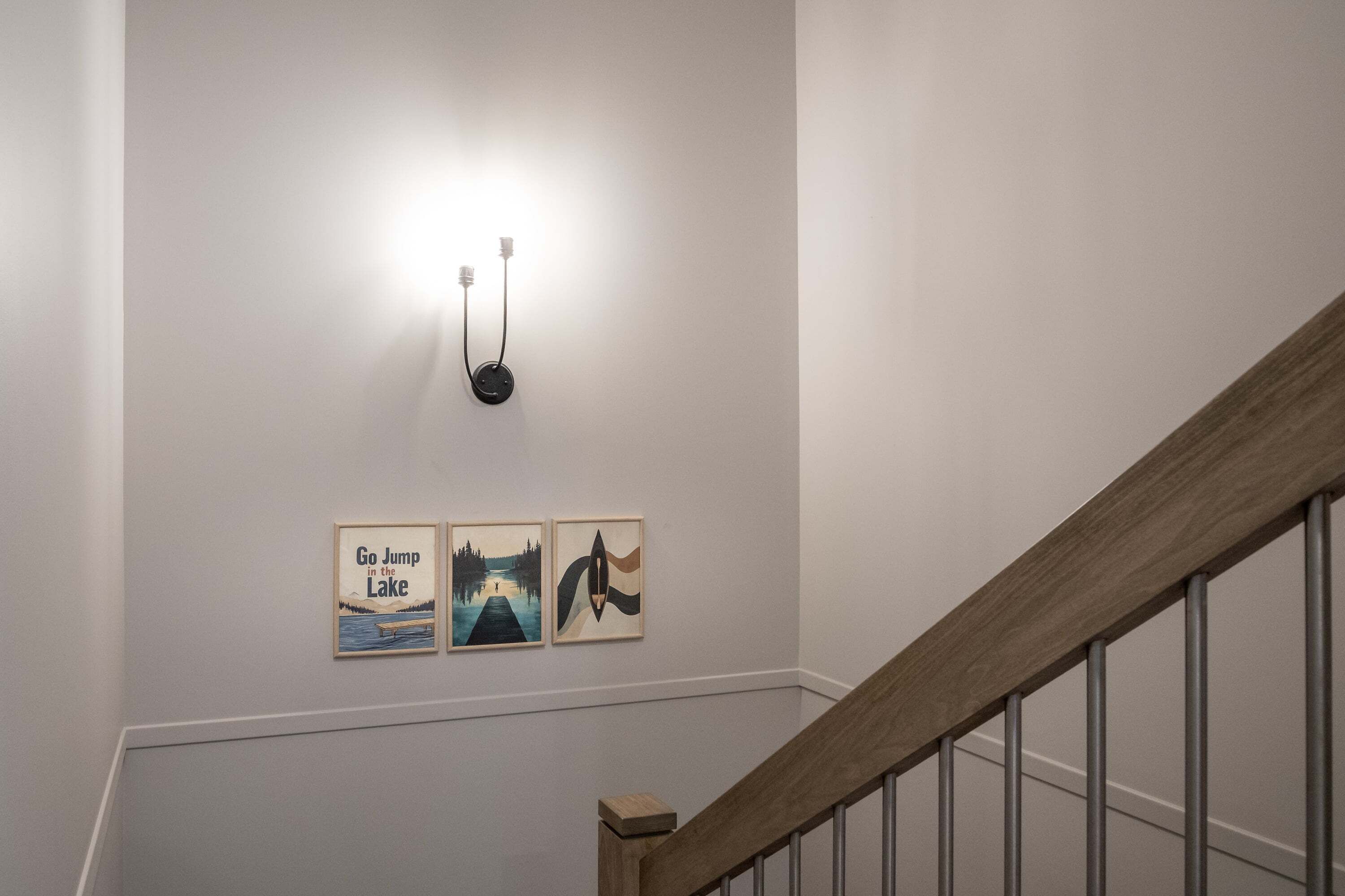 A staircase with a wooden handrail and metal balusters, leading to a landing with a wall-mounted light fixture and three framed artworks.