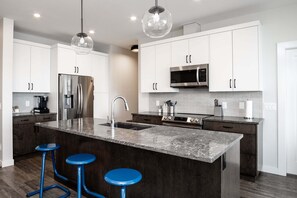A modern kitchen with white cabinets, stainless steel appliances, a marble island, and blue bar stools. - 3BR Kelowna townhome with lakeview, mountain views, deck, BBQ, Wi-Fi, and parking. (Kelowna)