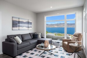A modern living room with a grey sectional sofa, a round coffee table, and a view of the sea through large windows. - 3BR Kelowna townhome with lakeview, mountain views, deck, BBQ, Wi-Fi, and parking. (Kelowna)