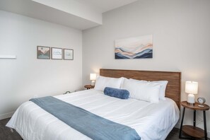 A hotel room with a large bed, two bedside tables, a nightstand, and a wall-mounted artwork. - 3BR Kelowna townhome with lakeview, mountain views, deck, BBQ, Wi-Fi, and parking. (Kelowna)