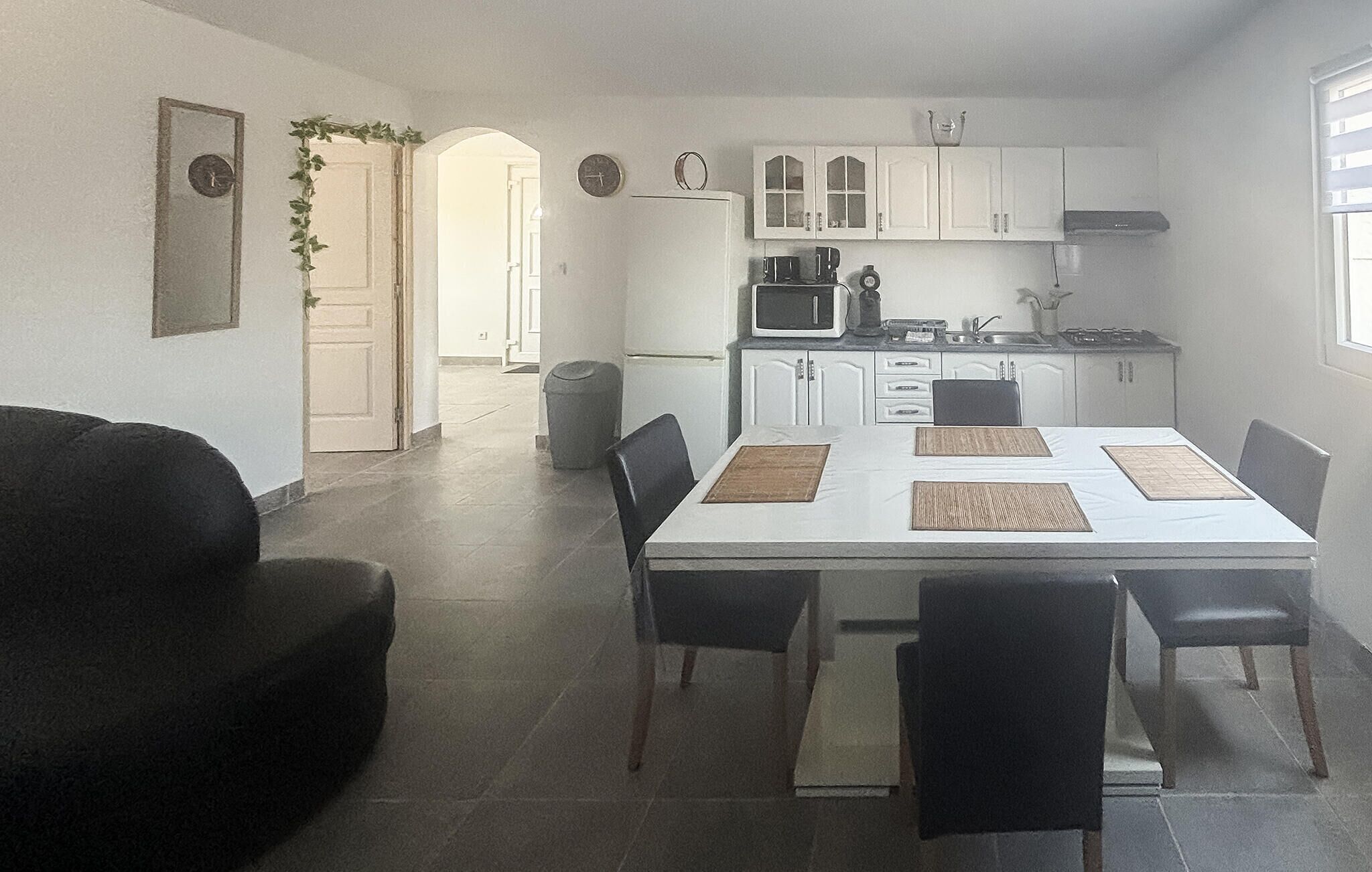Un comedor con una mesa blanca, sillas negras y un sofá negro. Se observa una cocina al fondo con armarios y electrodomésticos blancos.