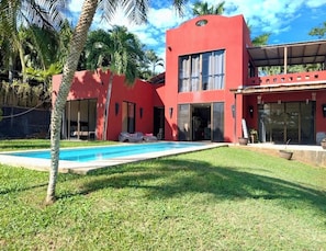 A red building with a swimming pool, palm trees, and a covered patio area. - 3 bdm*3.5 bath*short walk to the beach*pool*quiet (Tamarindo)
