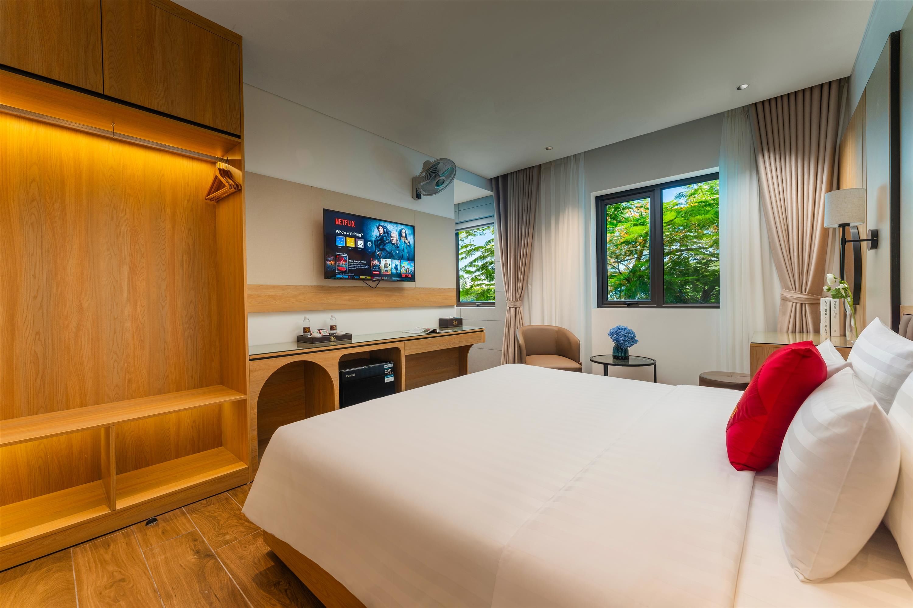 A hotel room with a large bed, a desk with a flat-screen TV, a seating area, and a window with a view of trees.