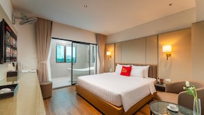 Quarto de hotel com cama, escrivaninha, cadeira, mesa de vidro, luminária e vista da cidade.