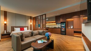 Quarto de hotel moderno com sofá, mesa de centro pequena, cama com travesseiro vermelho e cozinha equipada com armários de madeira e geladeira.