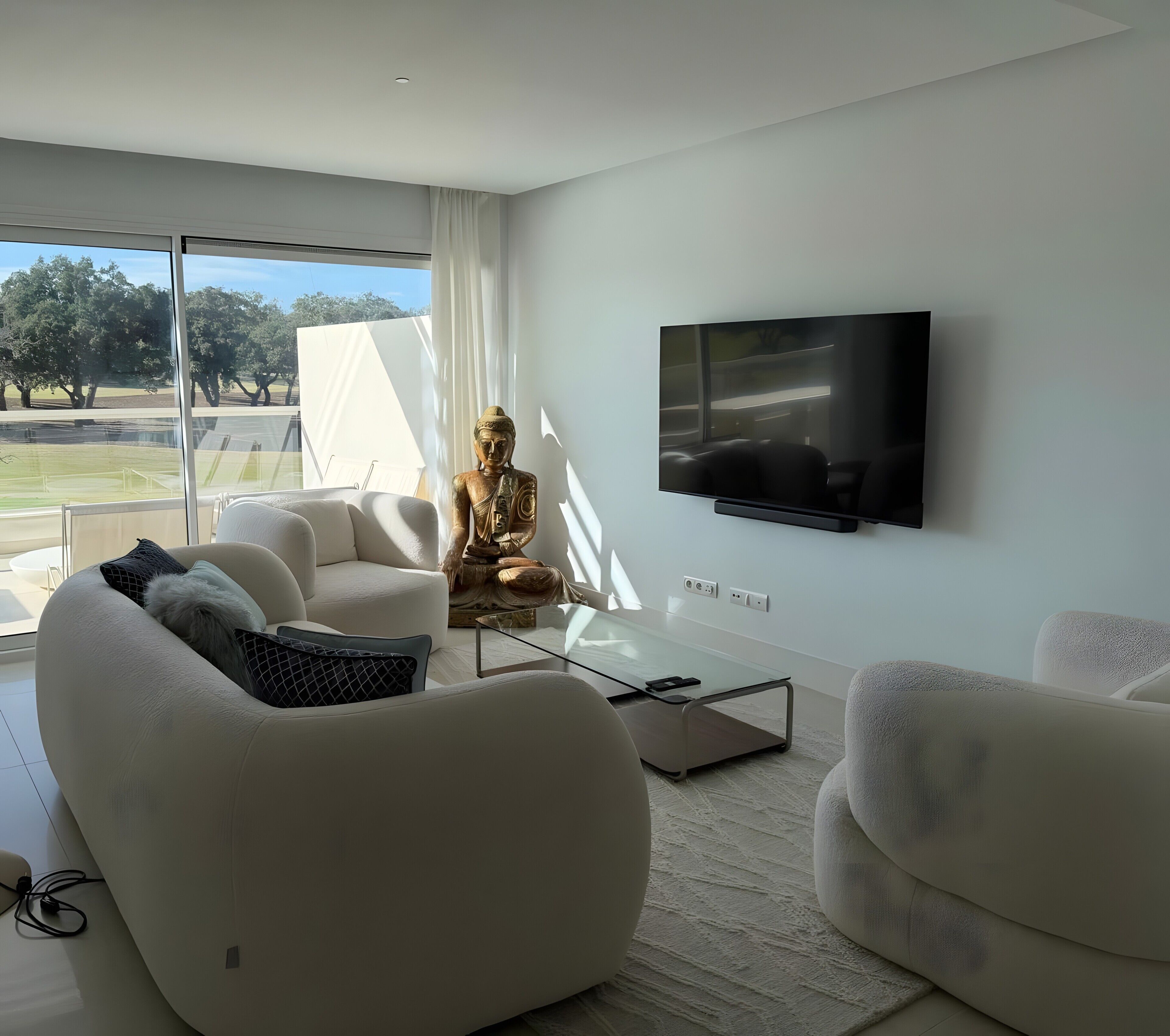 Una sala de estar moderna con televisor de pantalla plana, sofás blancos, una mesa de centro de vidrio y una estatua de Buda.