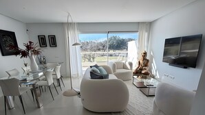A modern living room with a glass dining table, white sofas, and a large window offering a view of trees and a building. - Holiday Home 'Beautiful House in Emerald Green' with Sea View, Wi-Fi and Air Conditioning (San Roque)