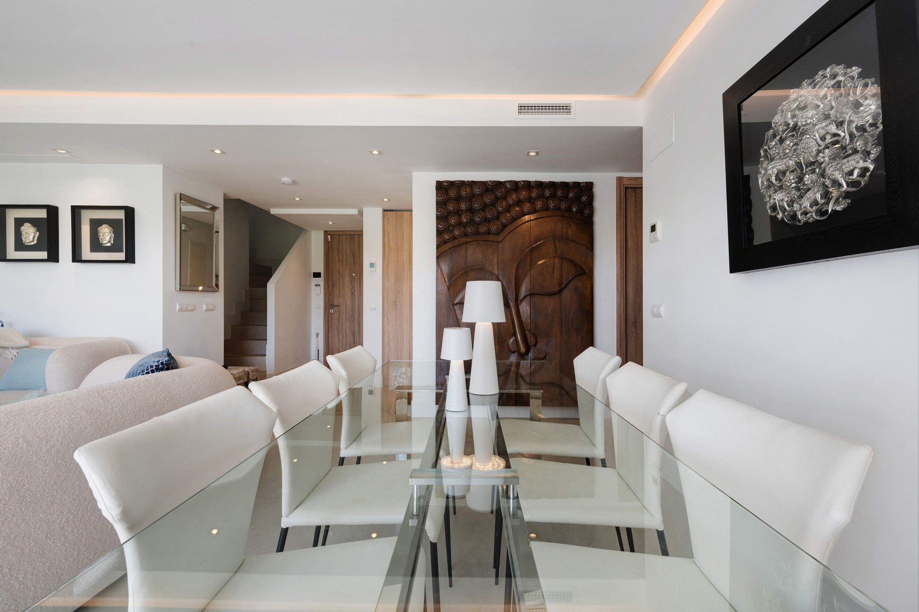 A modern dining area with a glass table and white chairs, a wooden door with a decorative panel, and a framed artwork above the door.