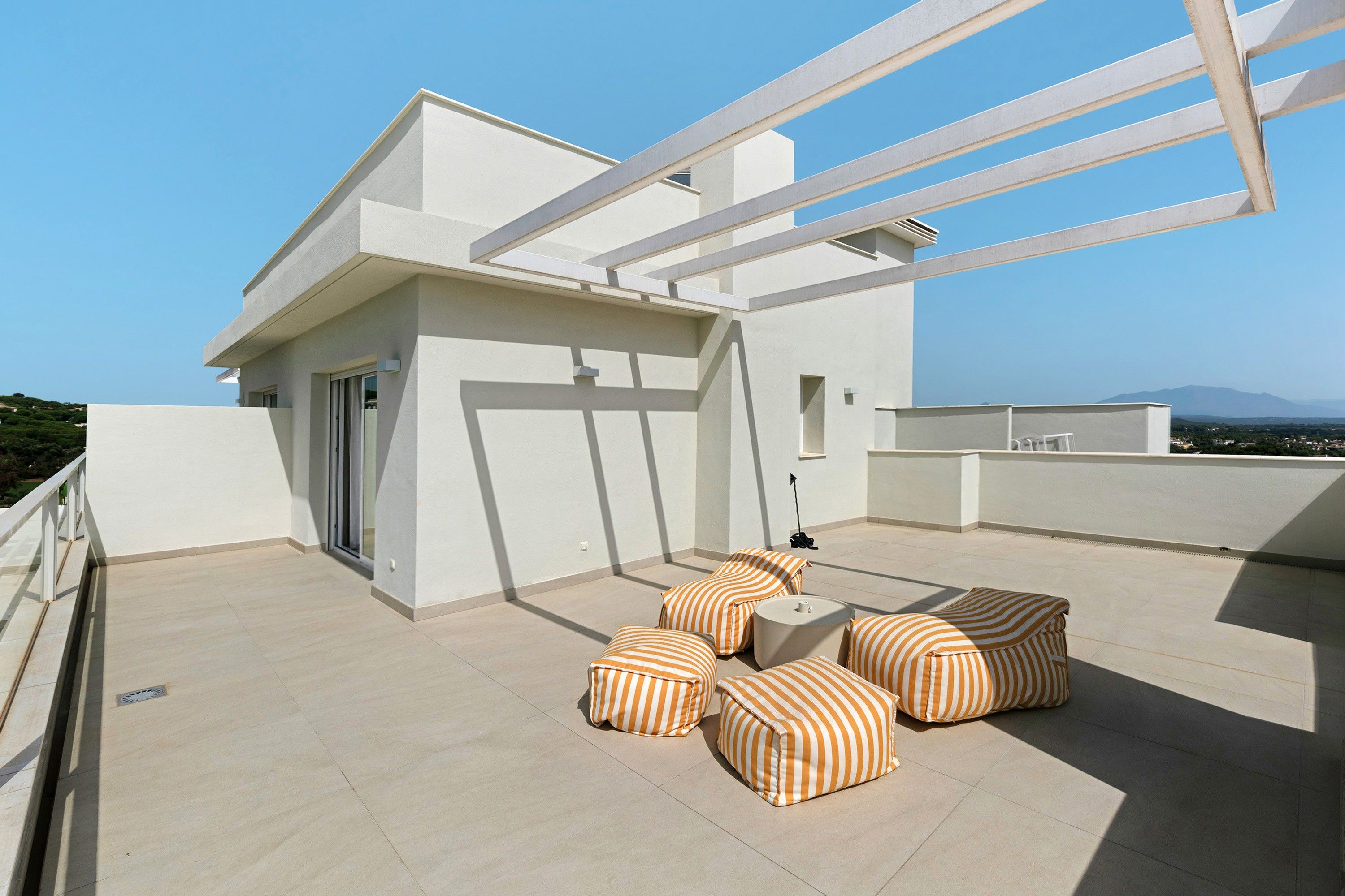 A modern outdoor terrace with a minimalist design, featuring a flat roof, a white railing, and a seating area with striped cushions.