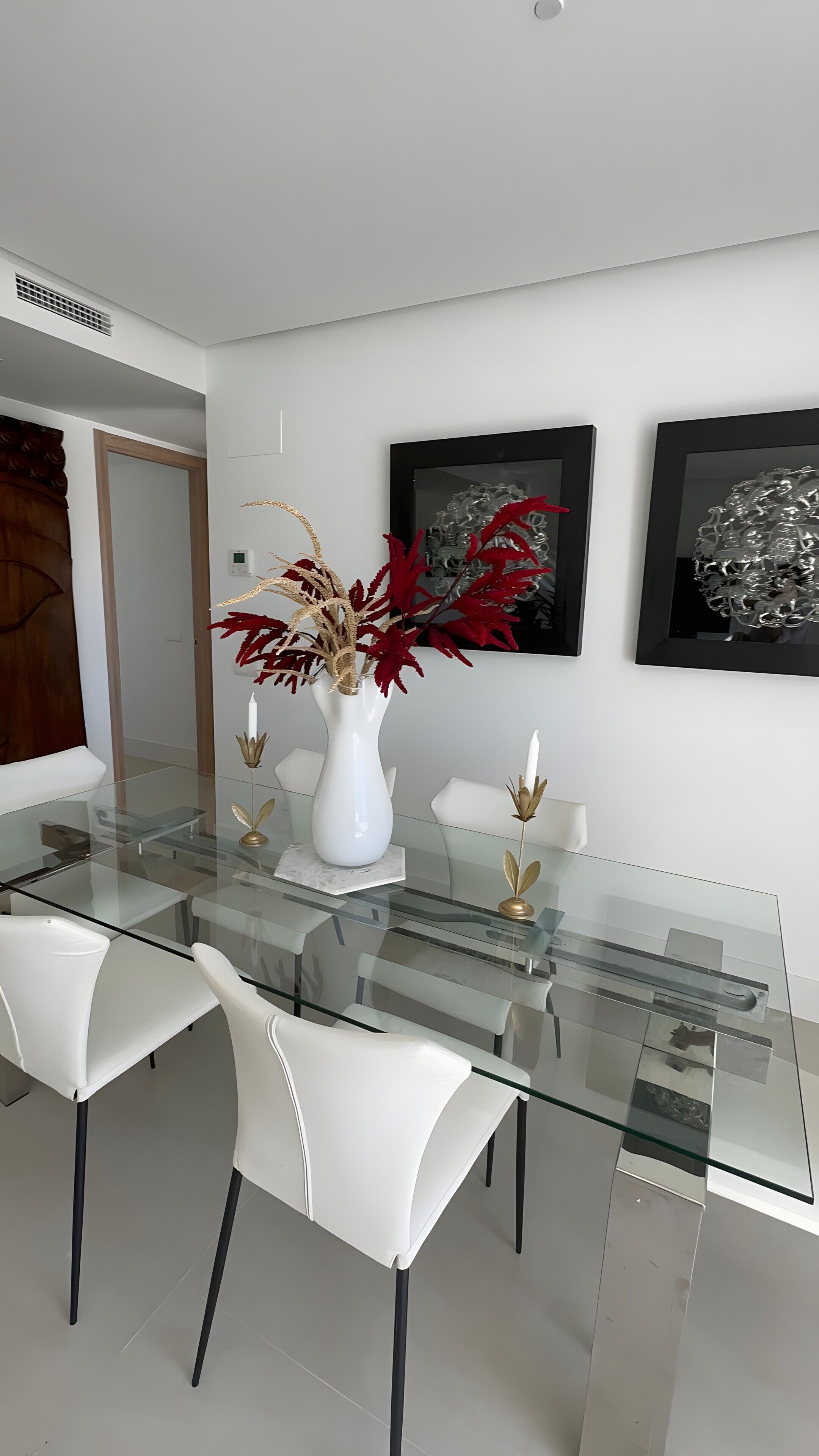 Un comedor moderno con una mesa de vidrio, sillas blancas y un florero con follaje rojo.
