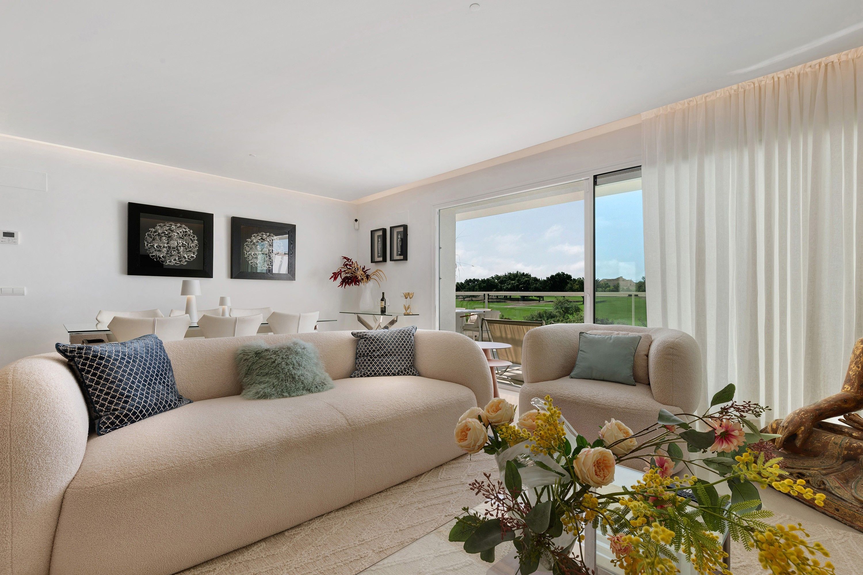 Una sala de estar moderna con un sofá color beige, dos sillones, una mesa de centro con flores y vistas a un paisaje verde a través de grandes ventanales.