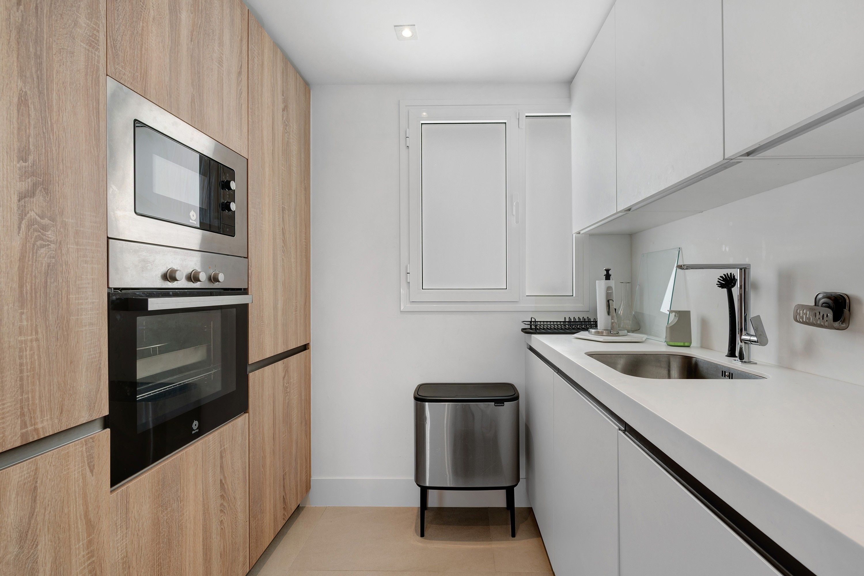 A modern kitchen with stainless steel appliances, a wooden cabinet, a white countertop, and a stainless steel trash bin.