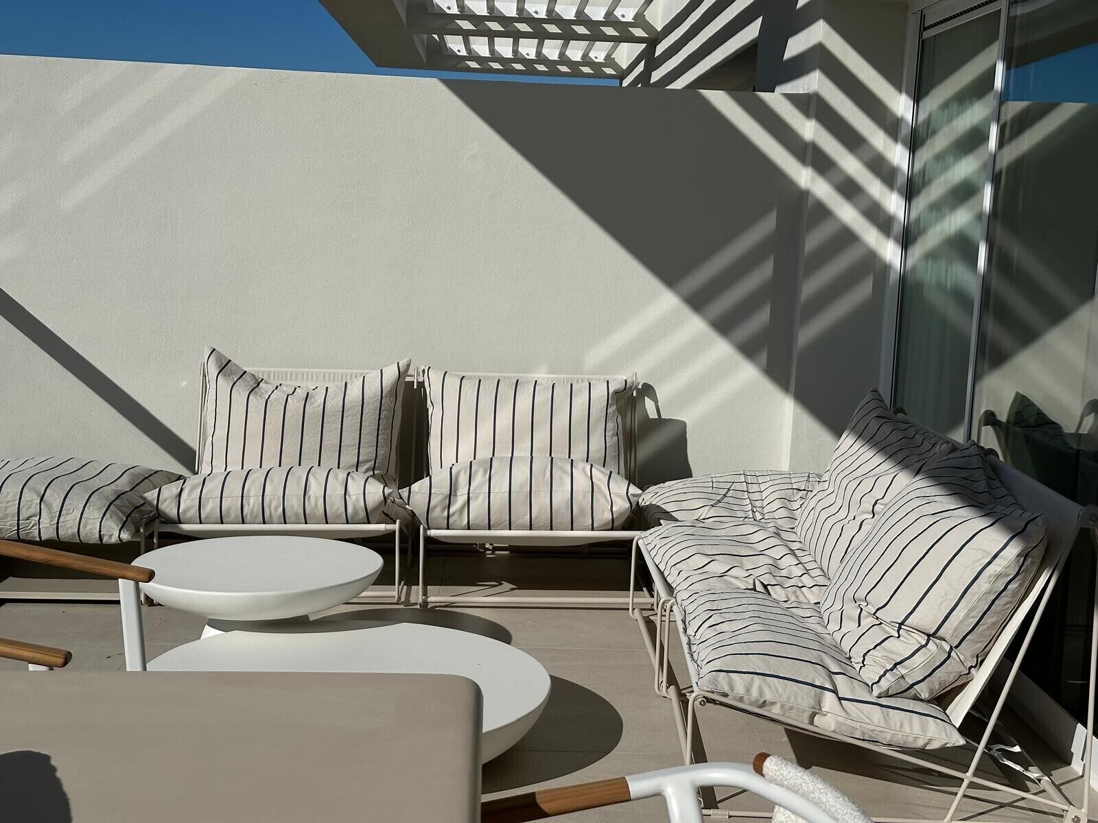 A sunlit outdoor seating area with a sofa and chairs, a round table, and a glass railing.