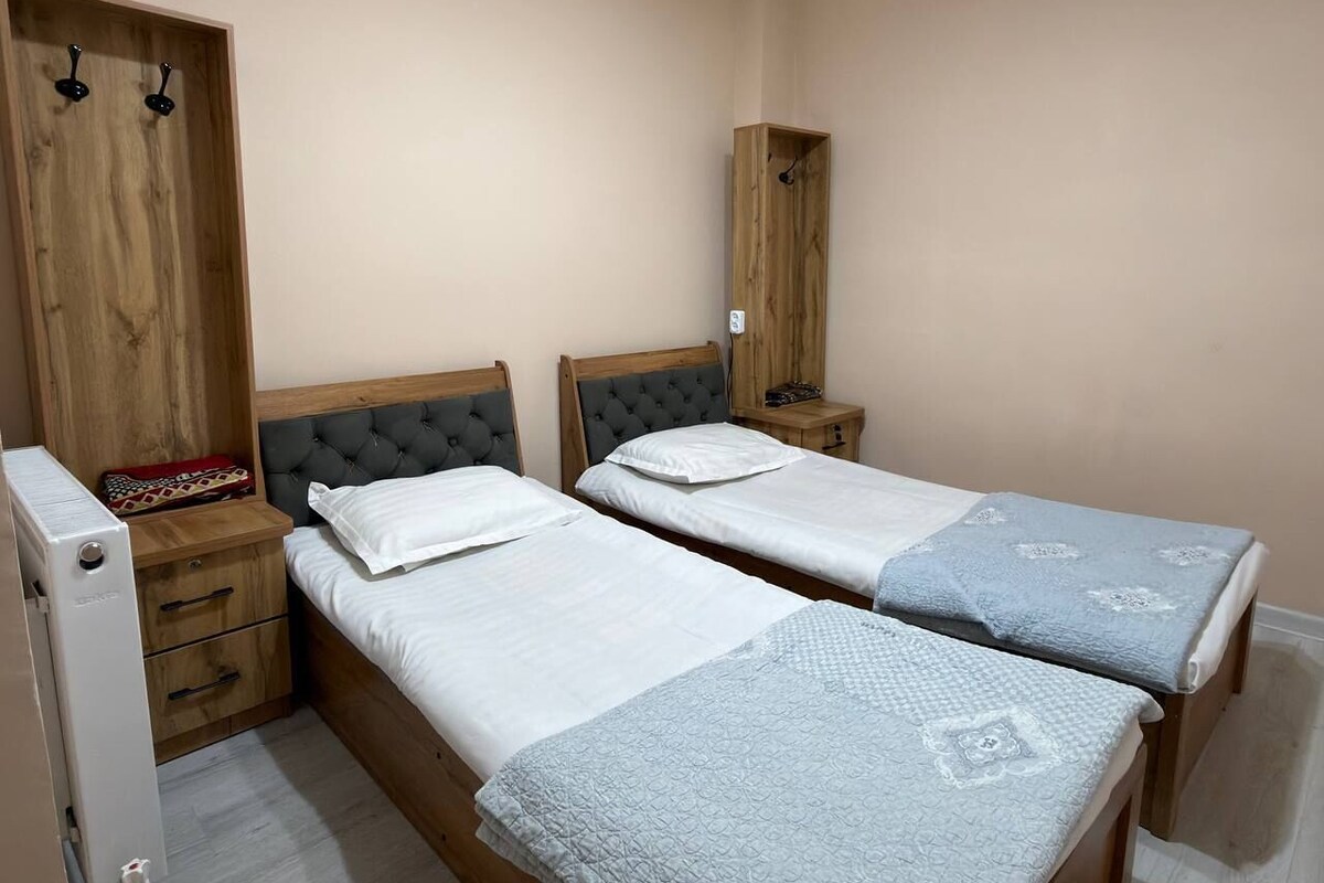 A bedroom with two beds, wooden headboards, a wardrobe, and a small bedside table with a lamp.
