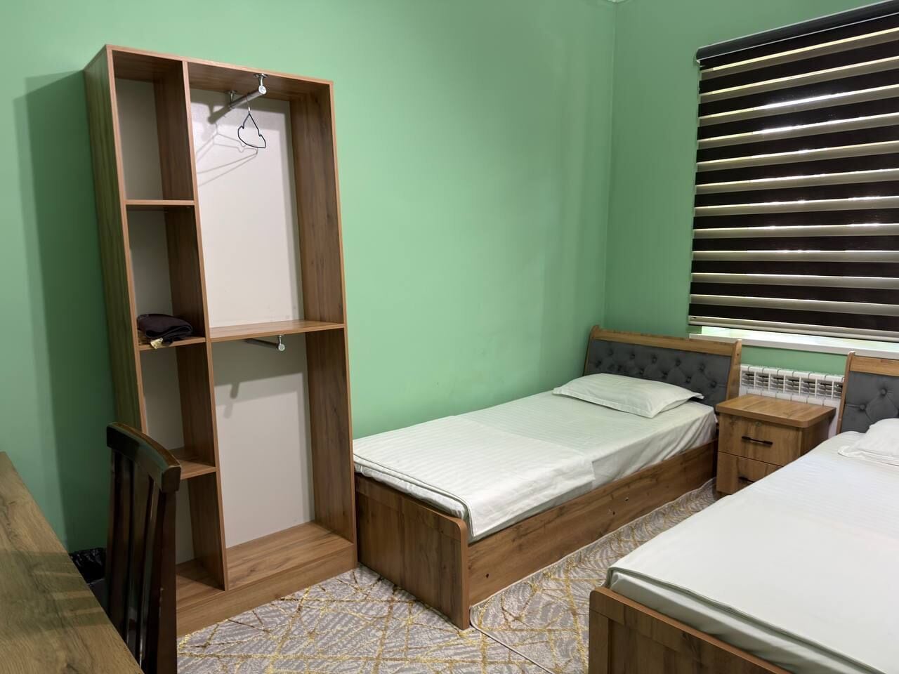 Une chambre avec un lit simple, une armoire en bois, une table de chevet et une fenêtre avec des stores.