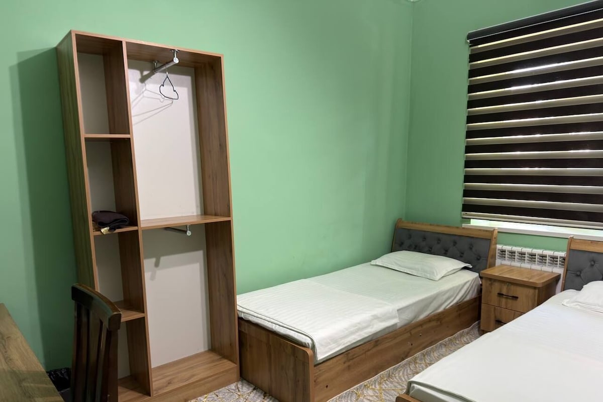 A bedroom with a single bed, a wooden wardrobe, a nightstand, and a window with blinds.
