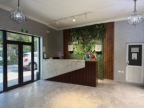 A reception area with a modern design, featuring a reception desk, a green wall with plants, and a view of parked cars outside. - Enjoy Hotel (Tashkent)