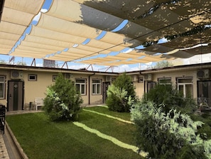 Un patio con un área de estar techada, un edificio con varias puertas y ventanas, y un césped con árboles.