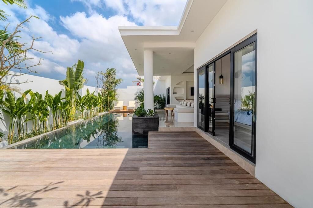 Un espacio exterior moderno con terraza de madera, piscina y un área acristalada con mobiliario.