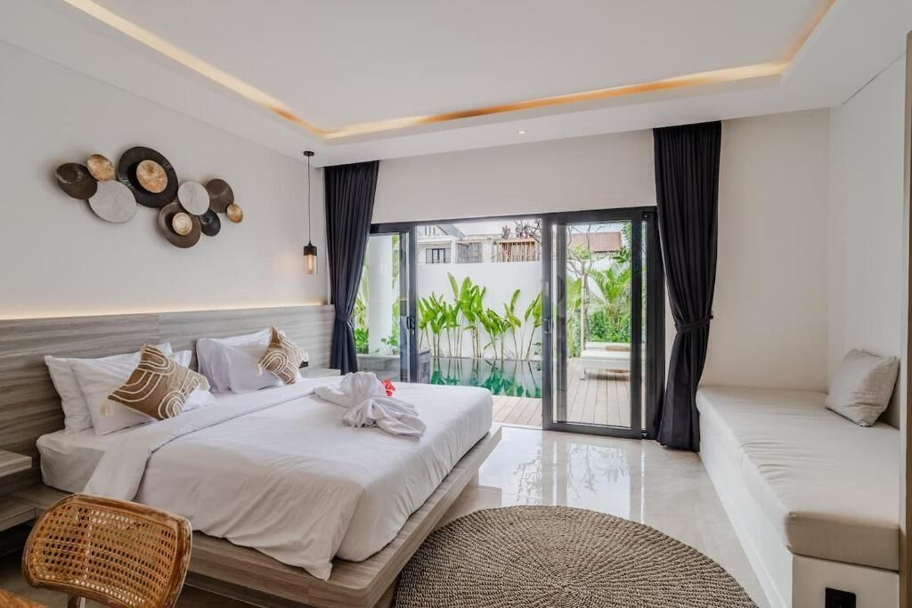Une chambre moderne avec un lit, un canapé, un fauteuil en rotin, un tapis tissé et une vue sur la verdure grâce à une porte-fenêtre coulissante.