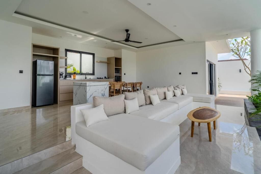 Een moderne woonkamer met een grote witte hoekbank, een kleine ronde houten tafel en een keuken op de achtergrond.