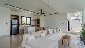 A modern living area with a large white sectional sofa, a small round wooden table, and a kitchen in the background. - Majys 4 Villa by Tasvan (Badung)