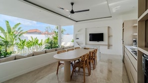 A modern kitchen with a flat-screen TV, a dining area with a large table and chairs, and a ceiling fan. - Majys 4 Villa by Tasvan (Badung)