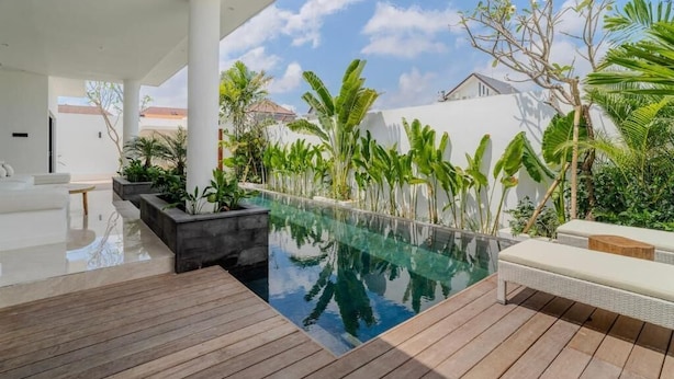 A modern outdoor pool area with a wooden deck, lounge chairs, and tropical plants. - Majys 4 Villa by Tasvan (Badung)