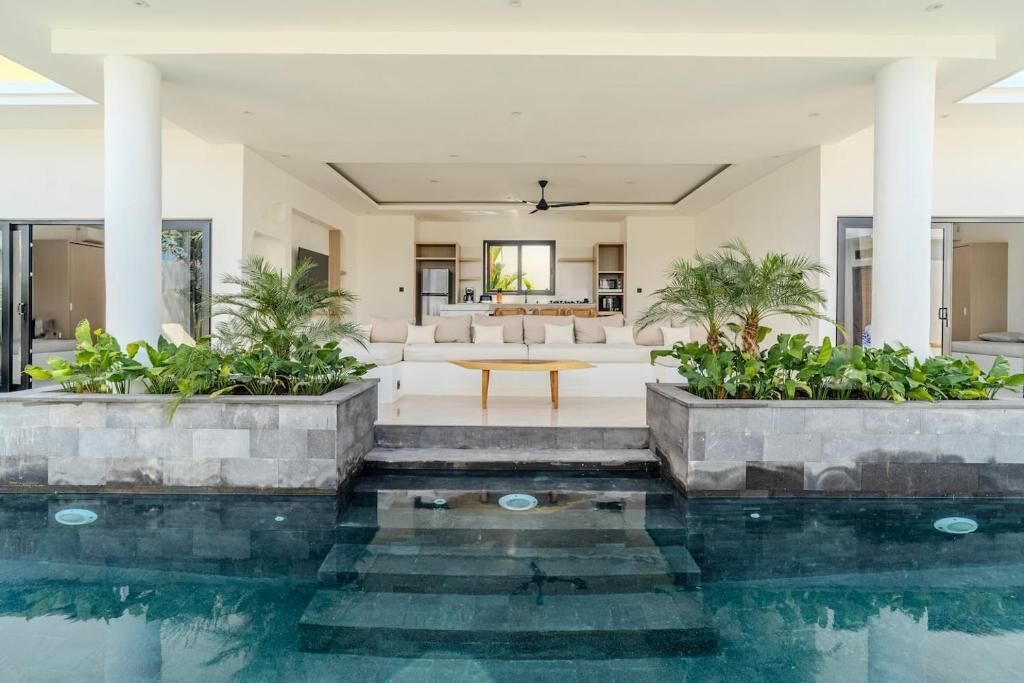 Un área de piscina interior moderna con zona de estar, ventilador de techo y plantas en macetas.