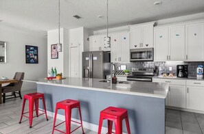 A modern kitchen with white cabinets, stainless steel appliances, and a central island with red stools. - 4BR Disney Getaway | Pool, Pirate & Castle Rooms (Davenport)