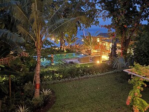 A pool area with a view of the sea, surrounded by tropical vegetation and a building with outdoor seating.