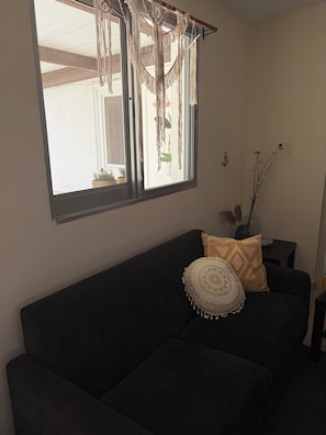 A dark-colored sofa with patterned cushions, a macrame wall hanging, and a window with a view of the outside. - Terra Olive Hideaway 3 bedroom villa (Pegeia)