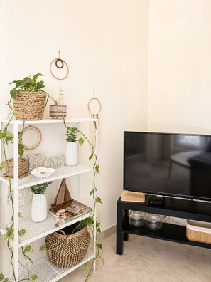 A living room with a white shelving unit, a black TV stand, and various decorative items including plants and woven baskets. - Terra Olive Hideaway 3 bedroom villa (Pegeia)