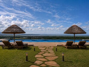 A poolside area with two lounge chairs under thatched umbrellas, a stone pathway, and a view of a body of water and distant landscape. - Africa Safari Manyara Migombani (Lake Manyara National Park)