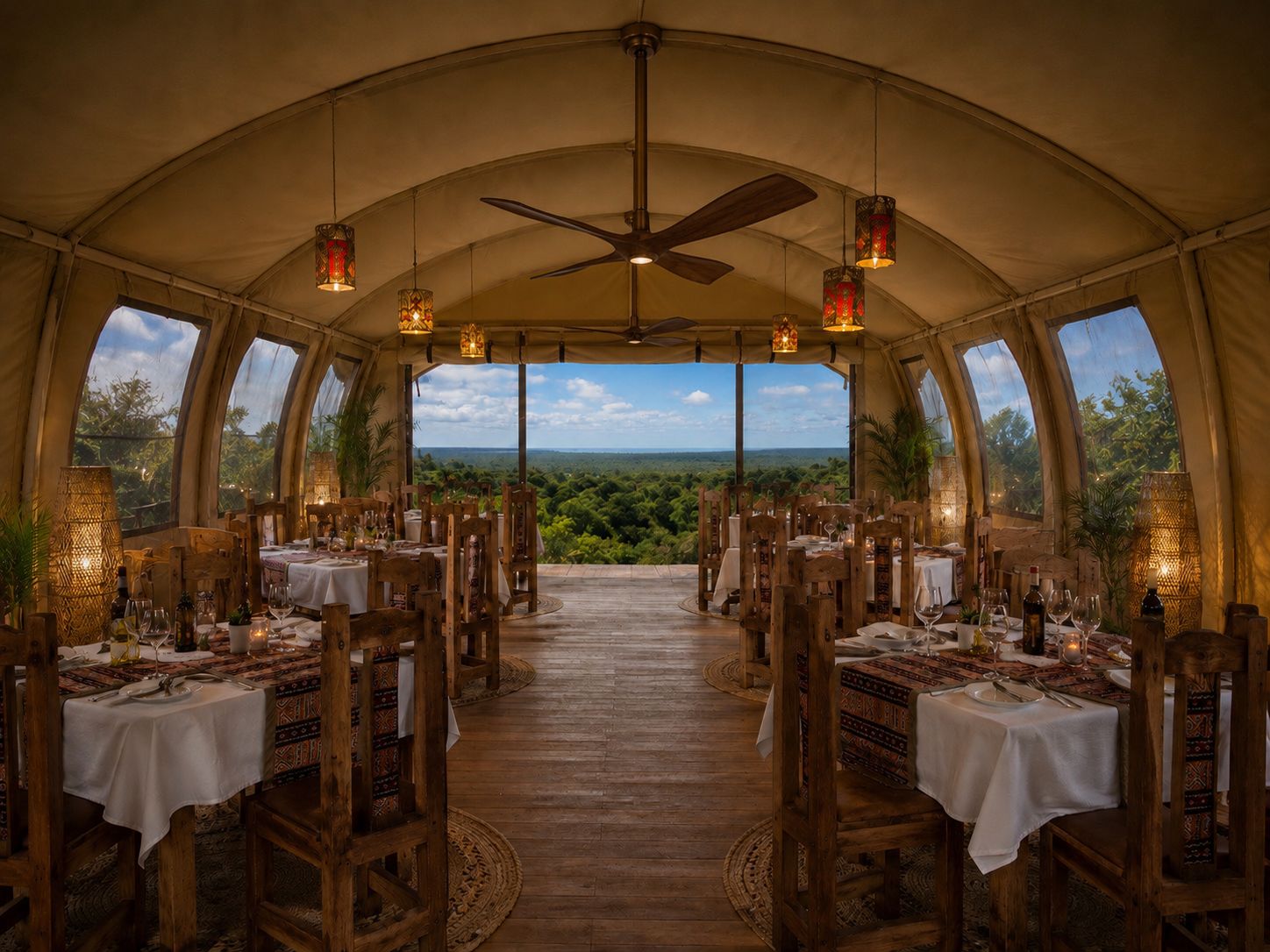 Ein Essbereich mit Zeltdach, Tischen, die für eine Mahlzeit gedeckt sind, einem Deckenventilator und großen Fenstern mit Blick auf die Landschaft.